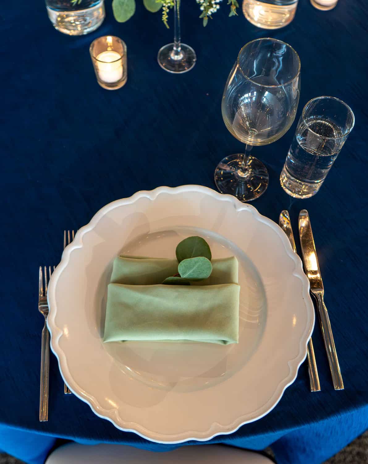 Table setting with a white plate, green napkin, and glassware on a blue tablecloth.