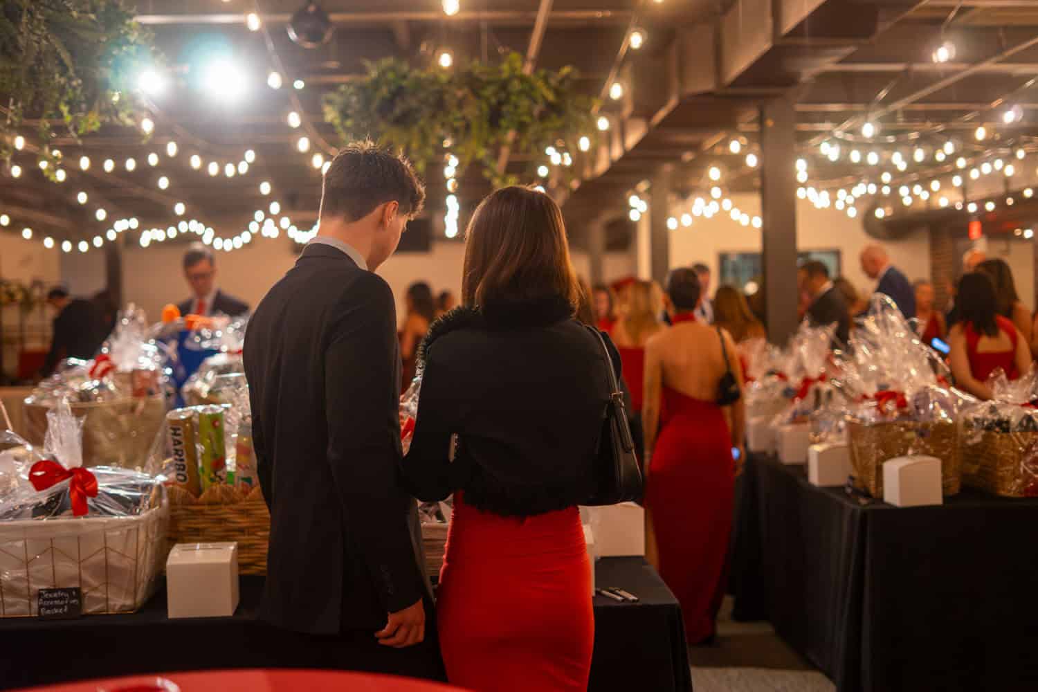 Couple in formal attire at festive holiday gathering with string lights.