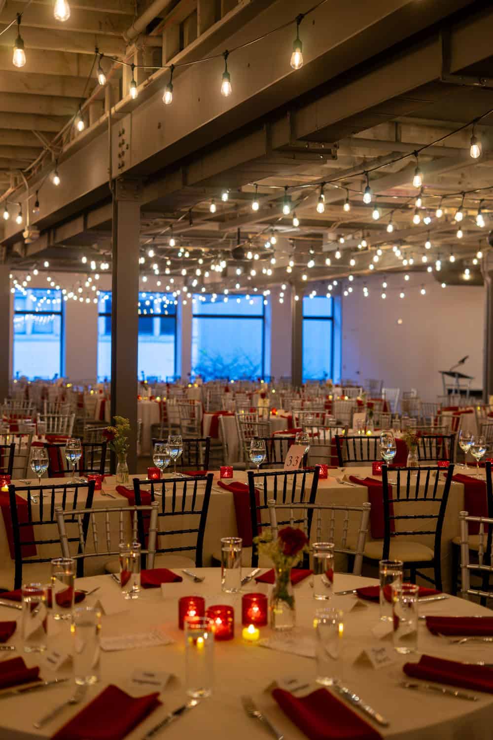 Indoor event space decorated with string lights and round tables for a celebration.