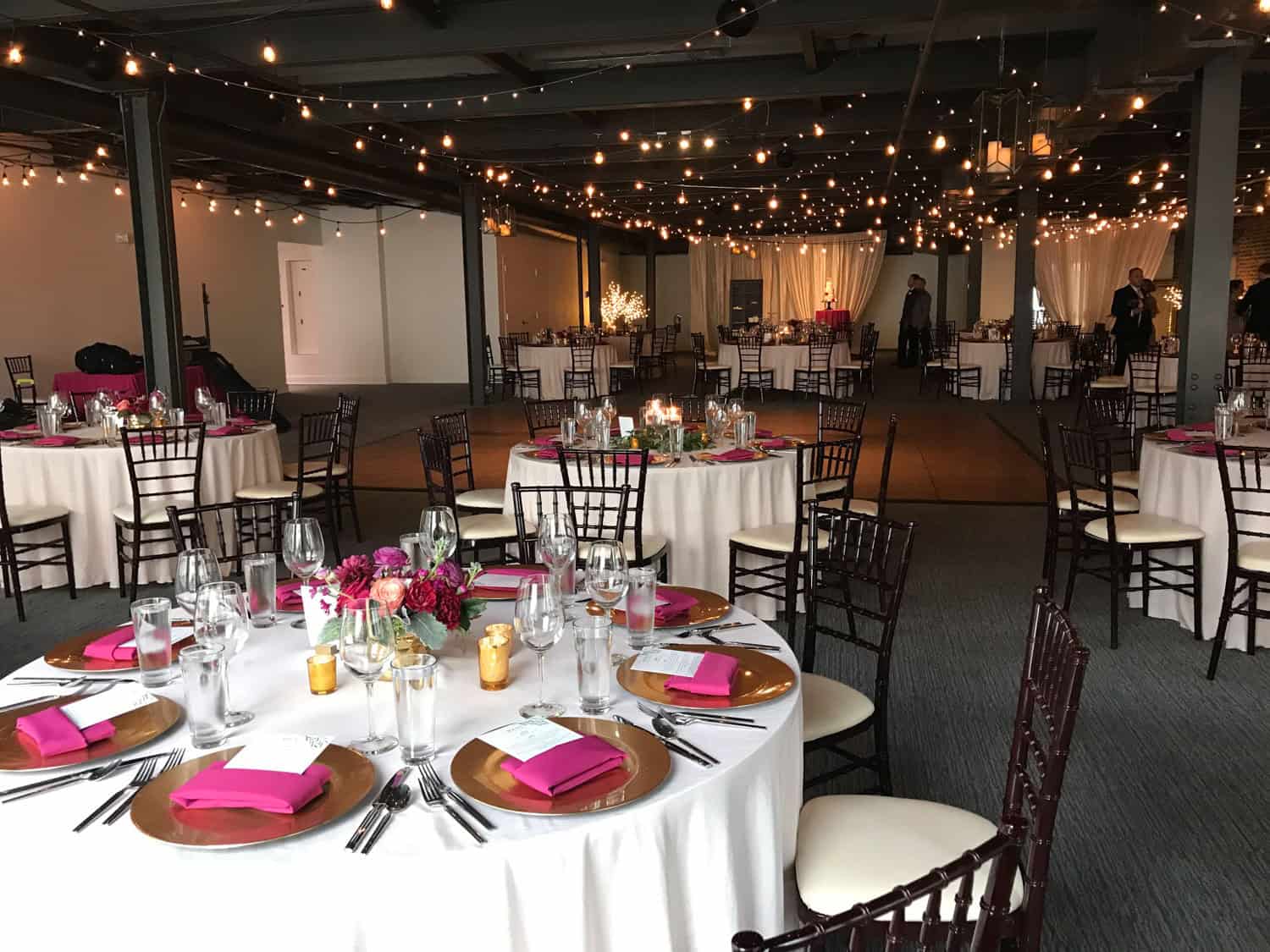 Decorated event space with round tables, string lights, and floral centerpieces.