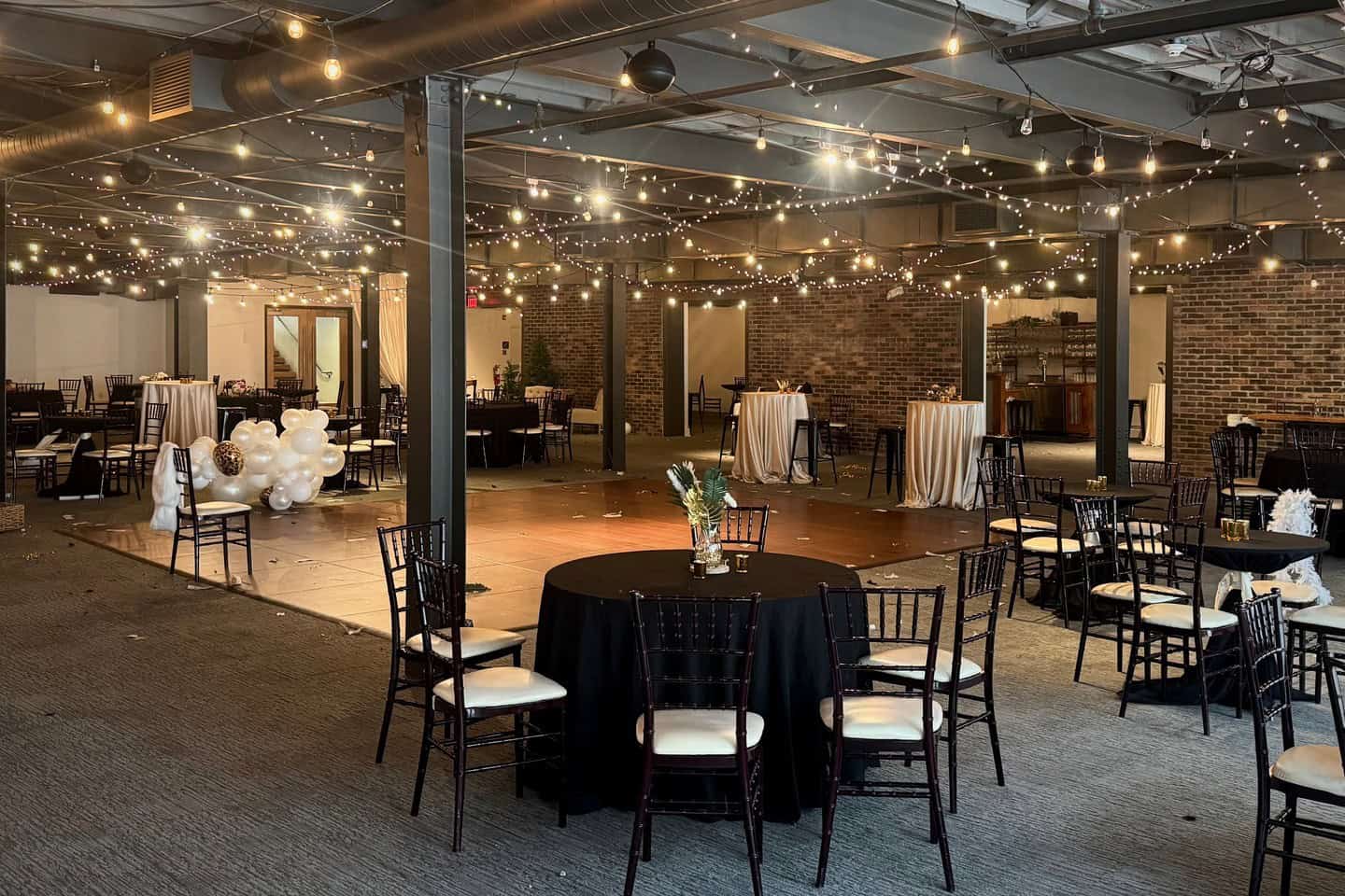Indoor event space decorated with string lights and round tables for a celebration.