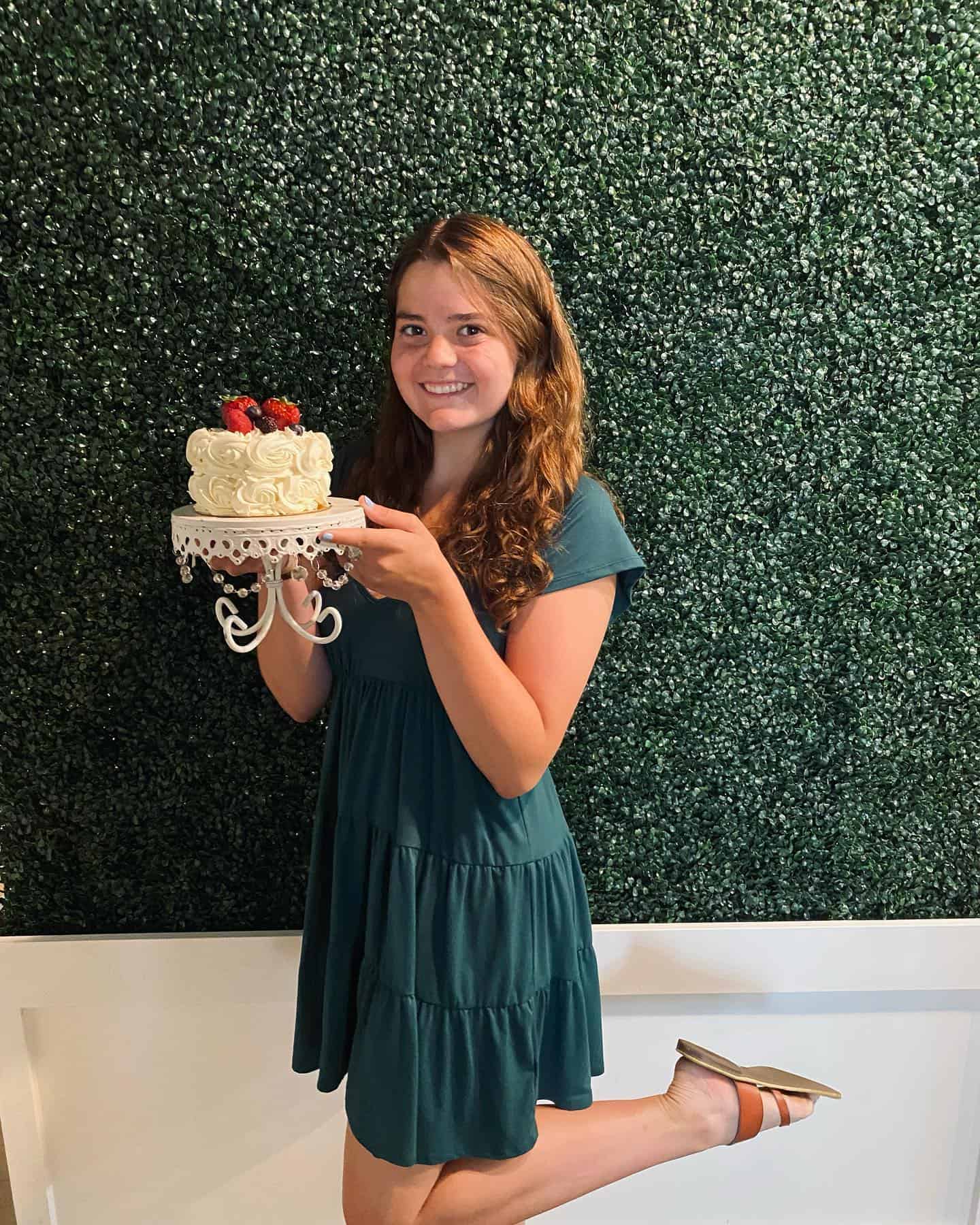 Young girl holding a birthday cake with strawberries in front of a green wall.