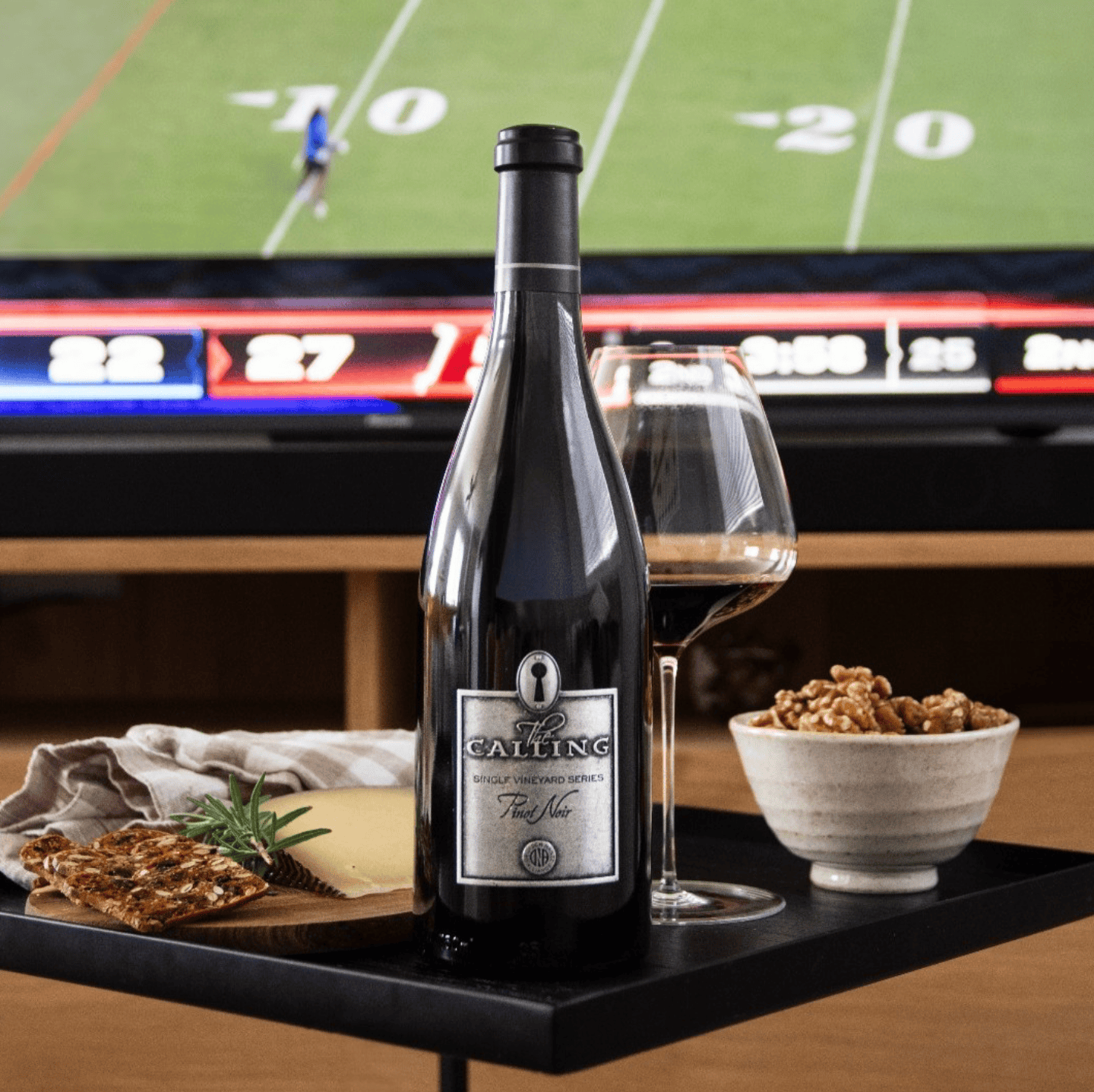 Wine bottle, glass, cheese, and snacks with football game in background.