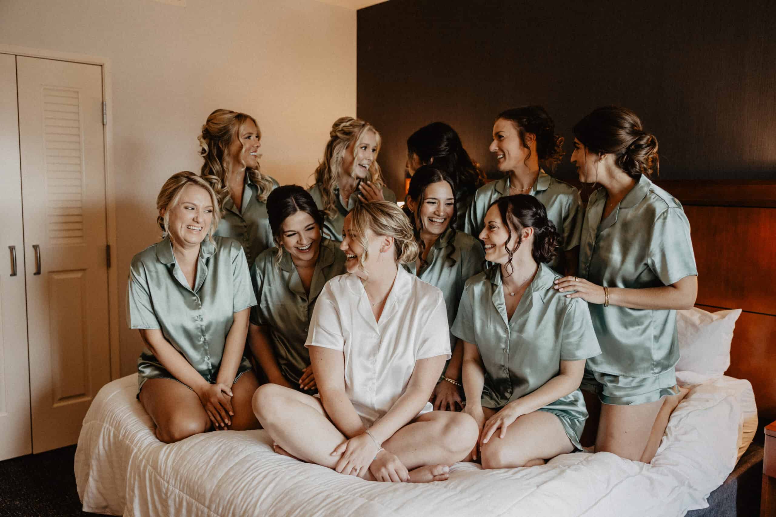 Group of bridesmaids in satin robes sitting on bed at SKY Armory hotel.