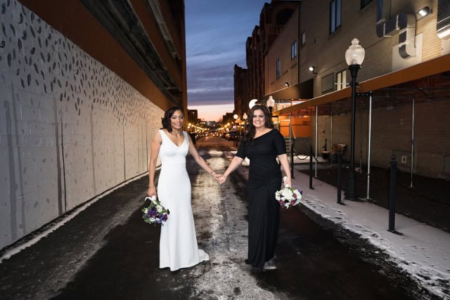 Two brides holding hands and holding bouquets in an urban alley at sunset.