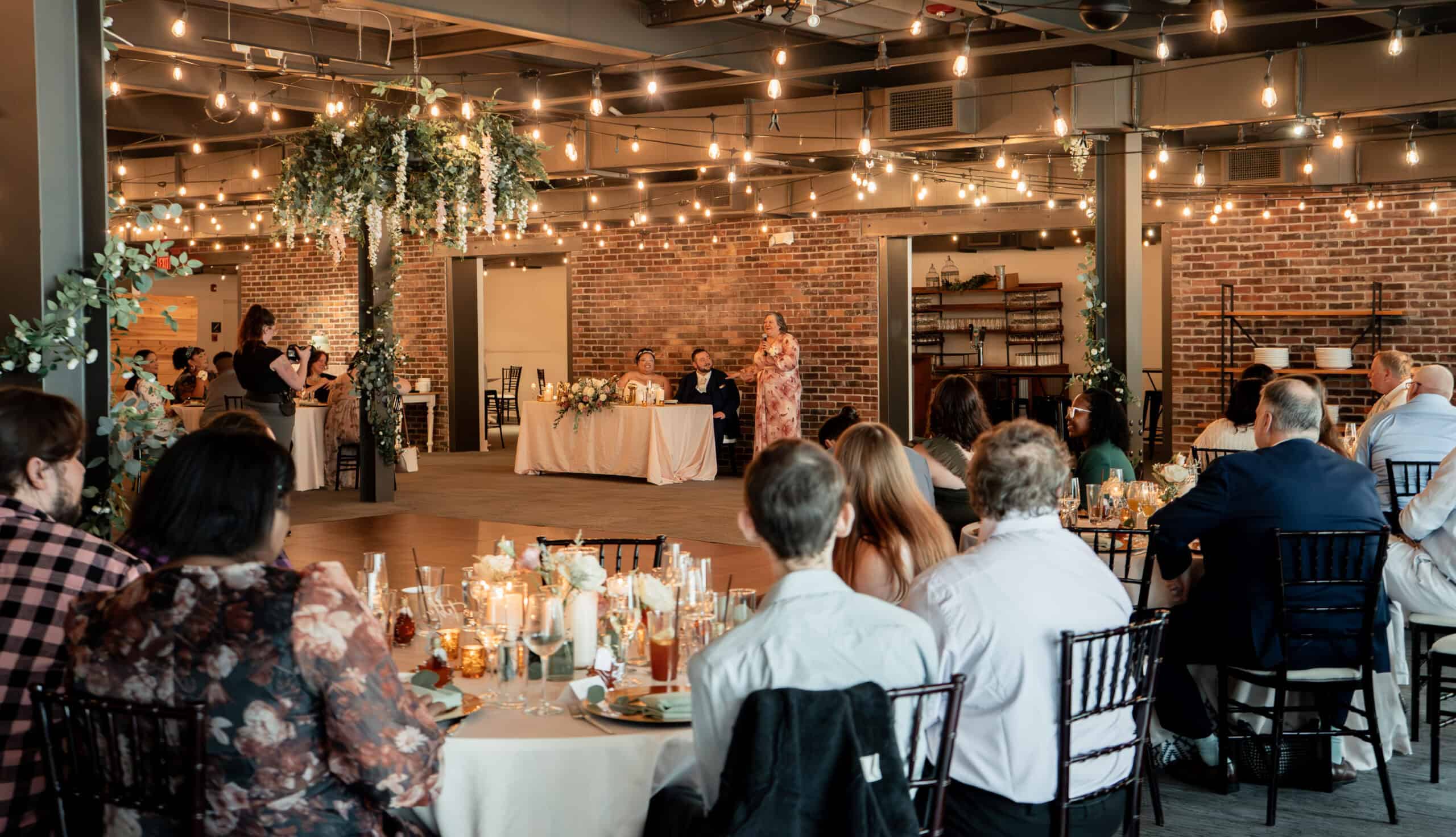 Elegant event space with string lights, brick walls, and seated guests at a celebration.