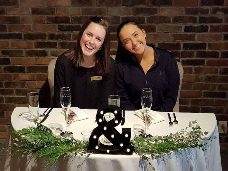 Two smiling women at a wedding reception table with greenery and candles.