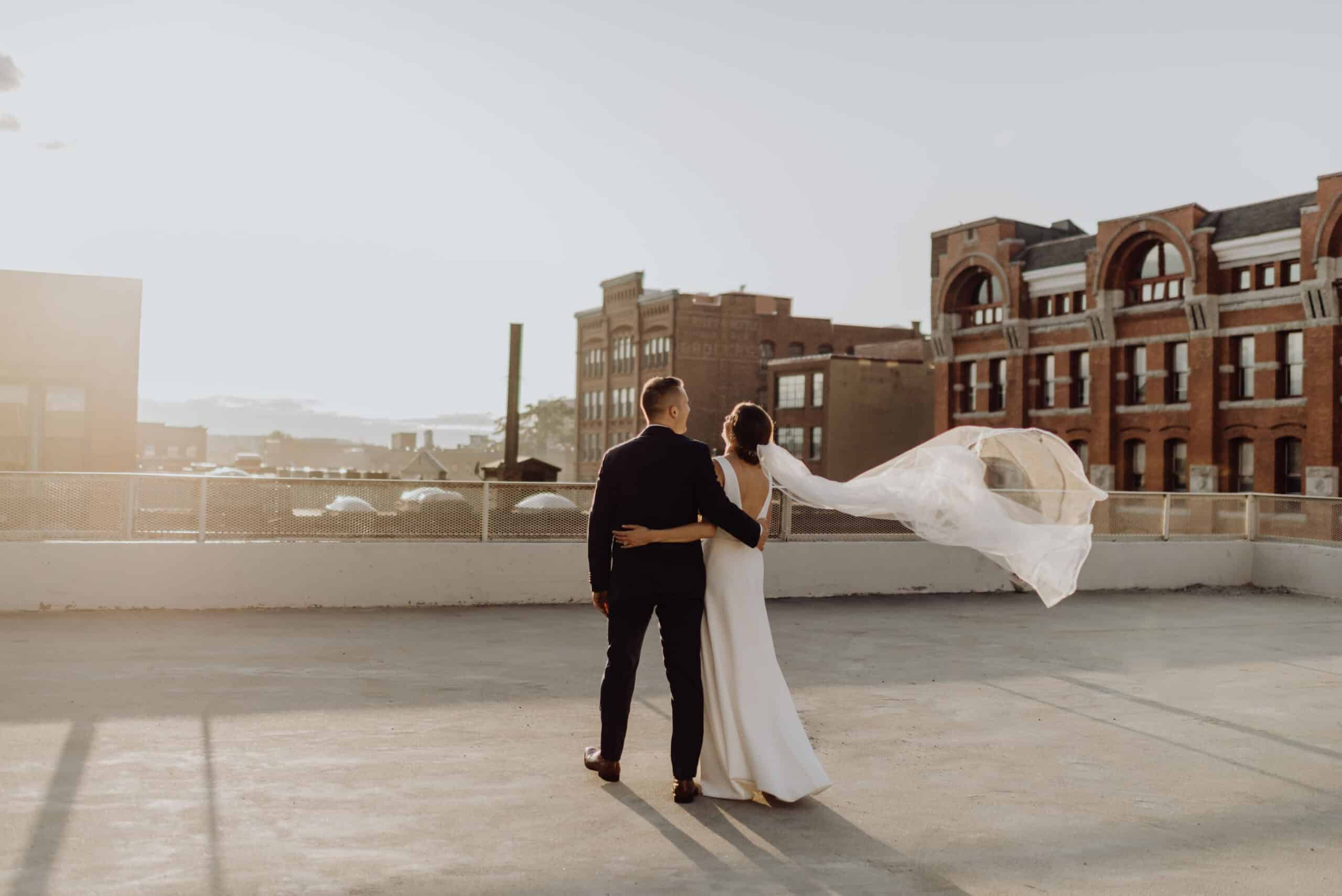 Bride and groom on rooftop with cityscape background during sunset.