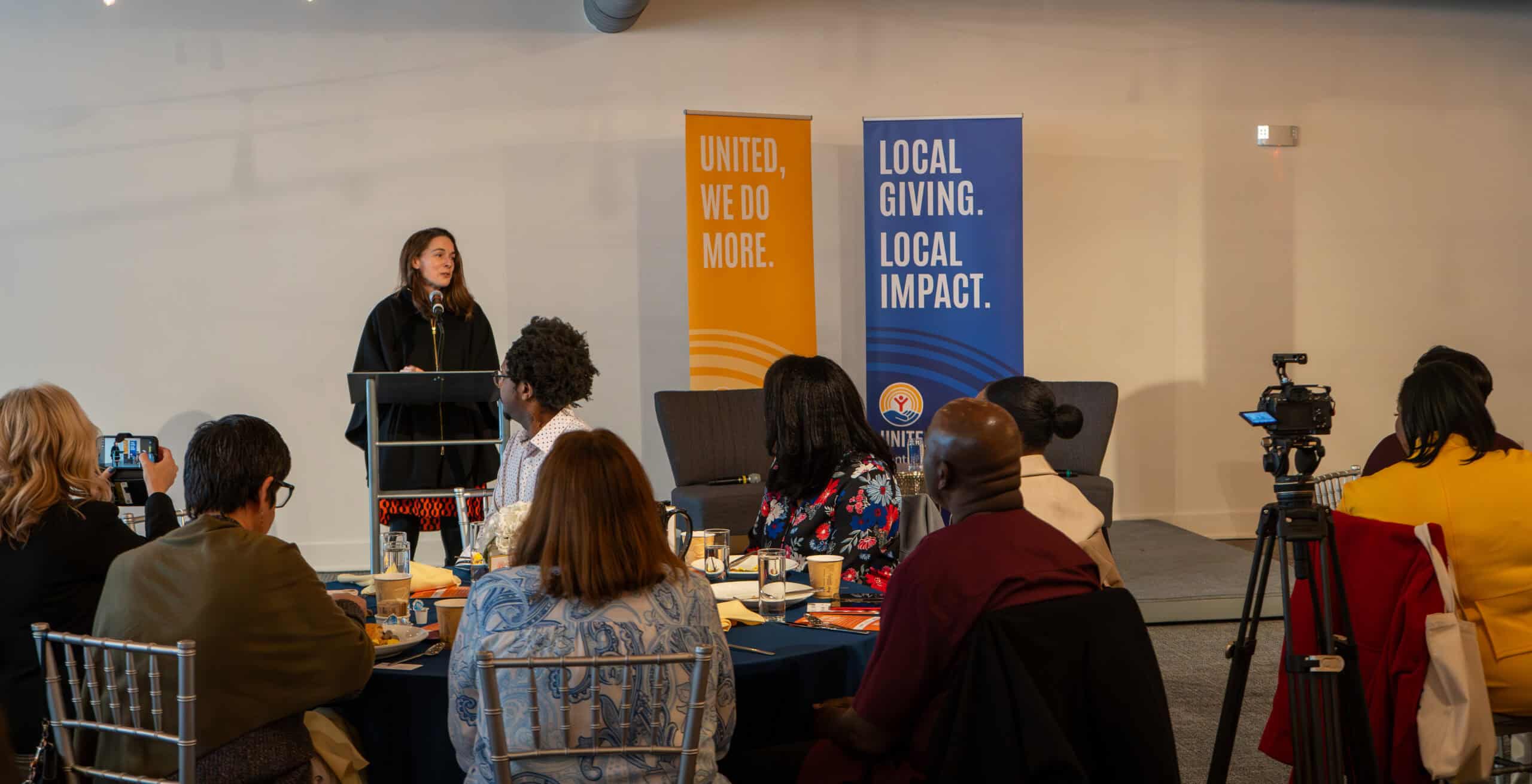 Speaker presenting at a corporate event with audience and banners in background.