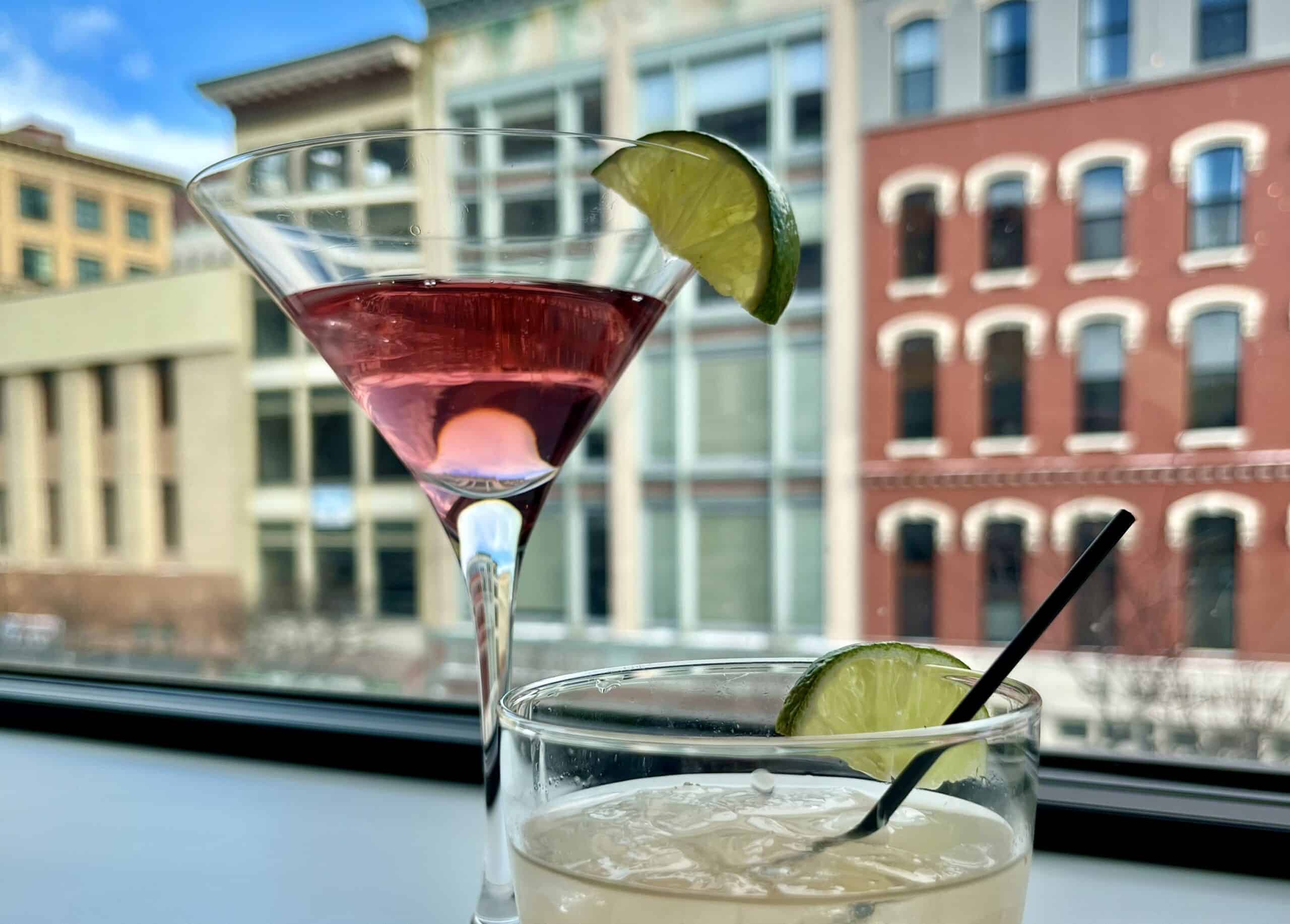Two cocktails with lime garnishes on a windowsill with city buildings in background.