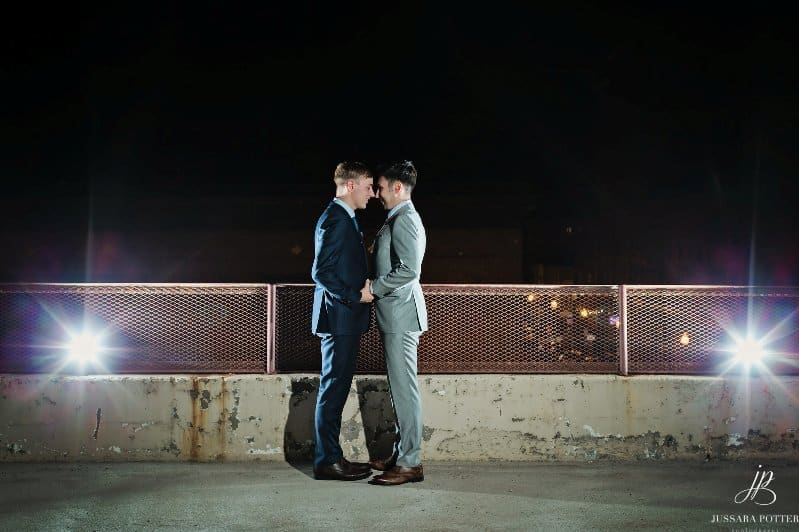 Two grooms in suits face each other on rooftop at night.