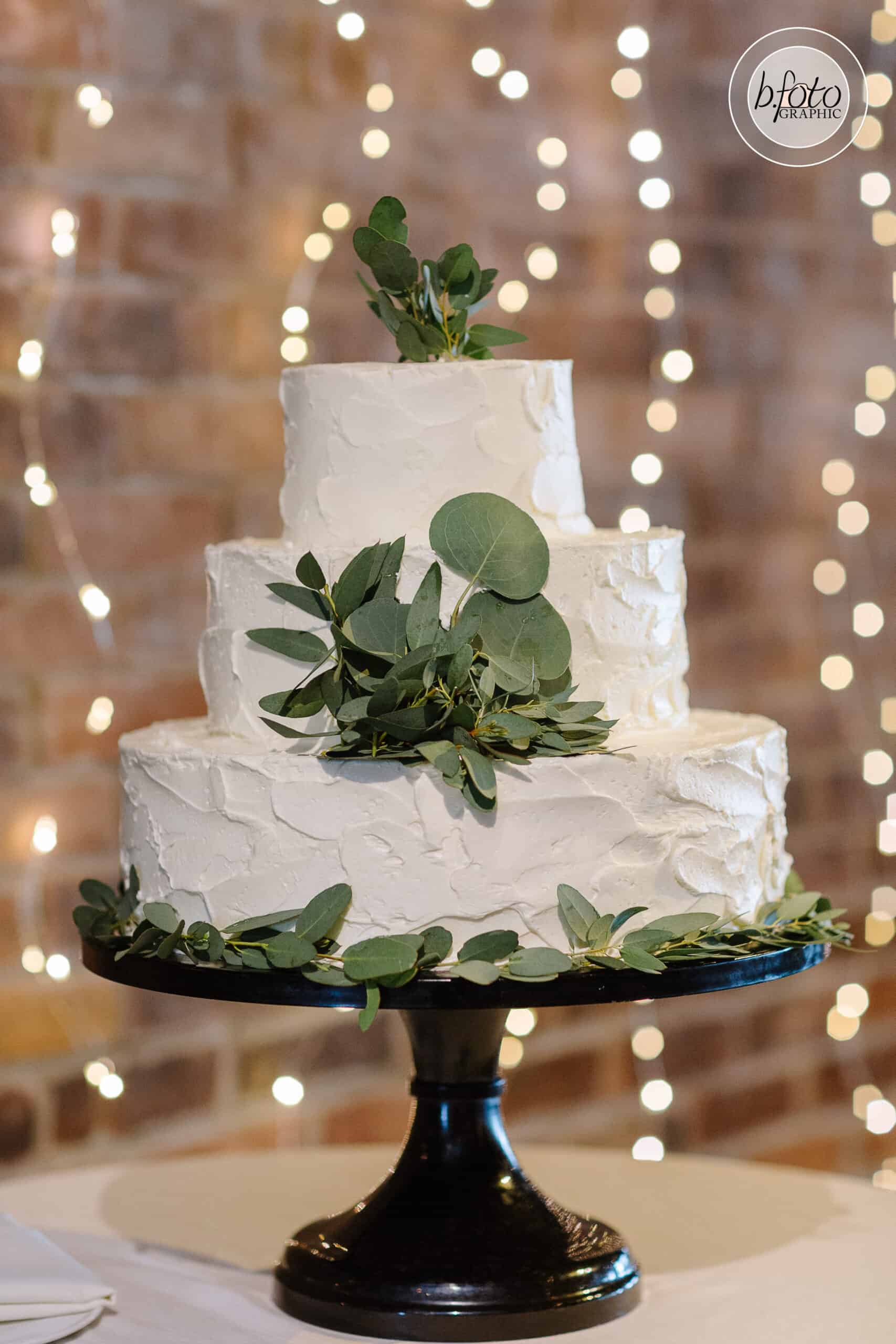 Three-tier white wedding cake decorated with fresh greenery on a black cake stand.
