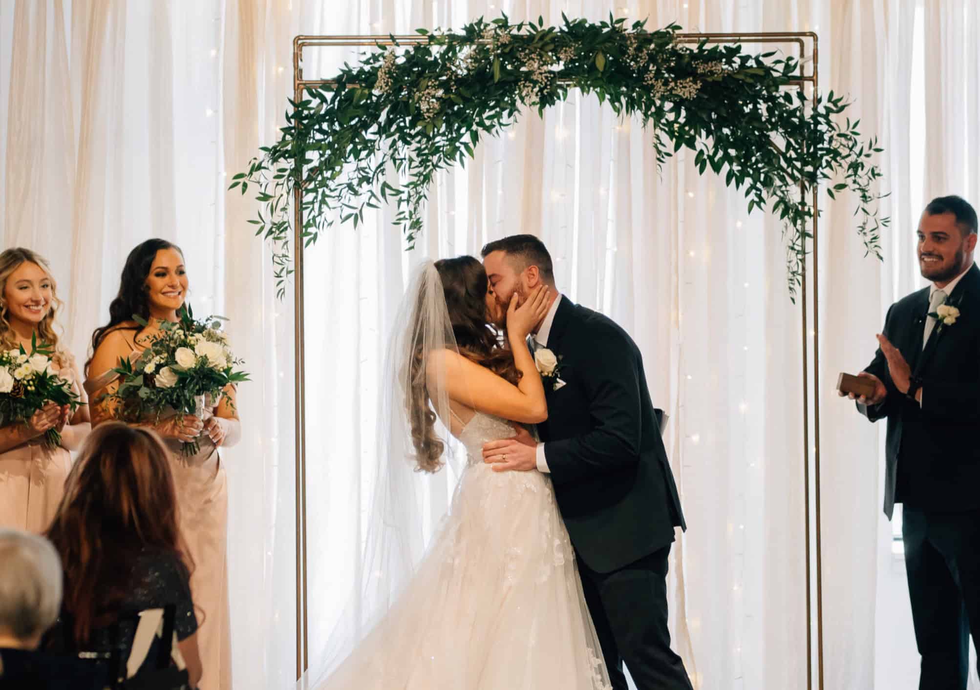 Bride and groom kiss during wedding ceremony at SKY Armory with floral arch.