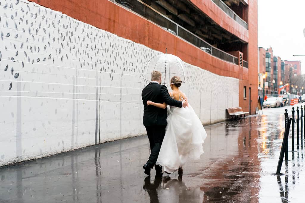 Bride and groom walking in the rain with umbrella in an urban setting.