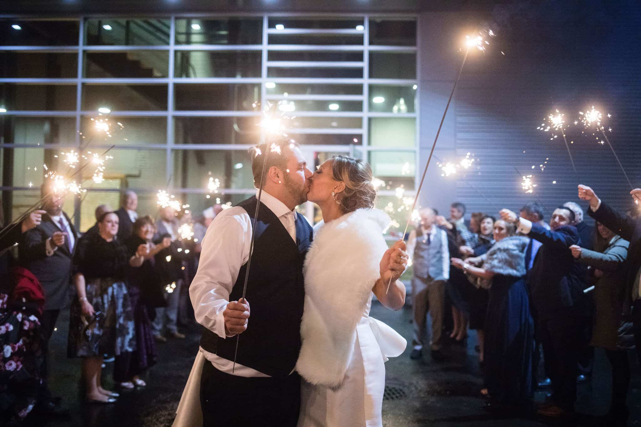 Couple kissing at wedding with sparklers and guests in background.