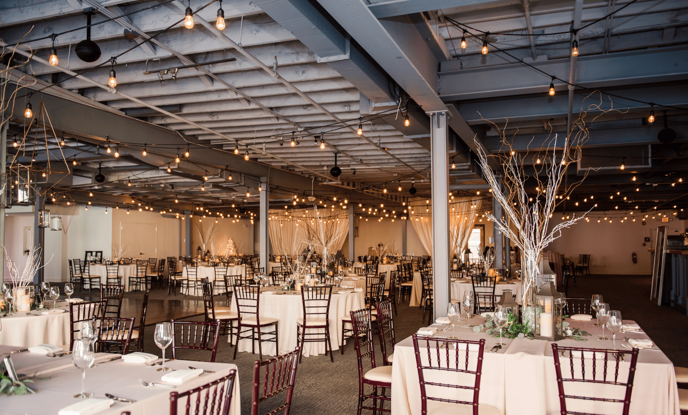 Indoor event space decorated with string lights and round tables for a celebration.