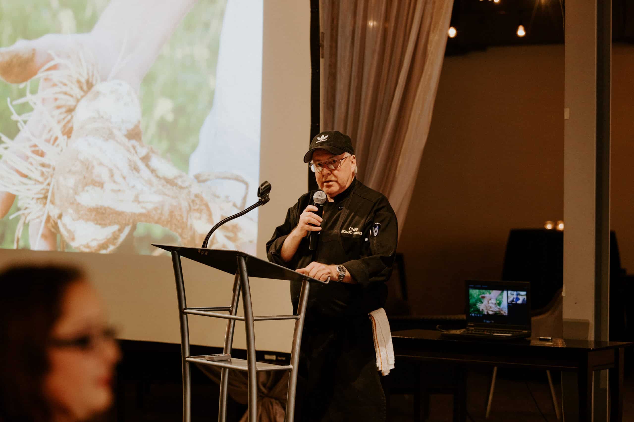 Chef speaking at SKY Armory event with presentation screen behind.