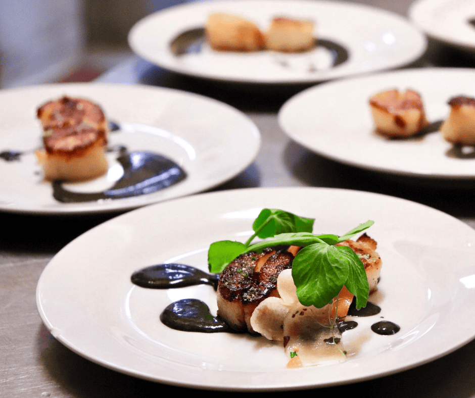 Elegant seared scallops garnished with basil and drizzled with black sauce on white plates.