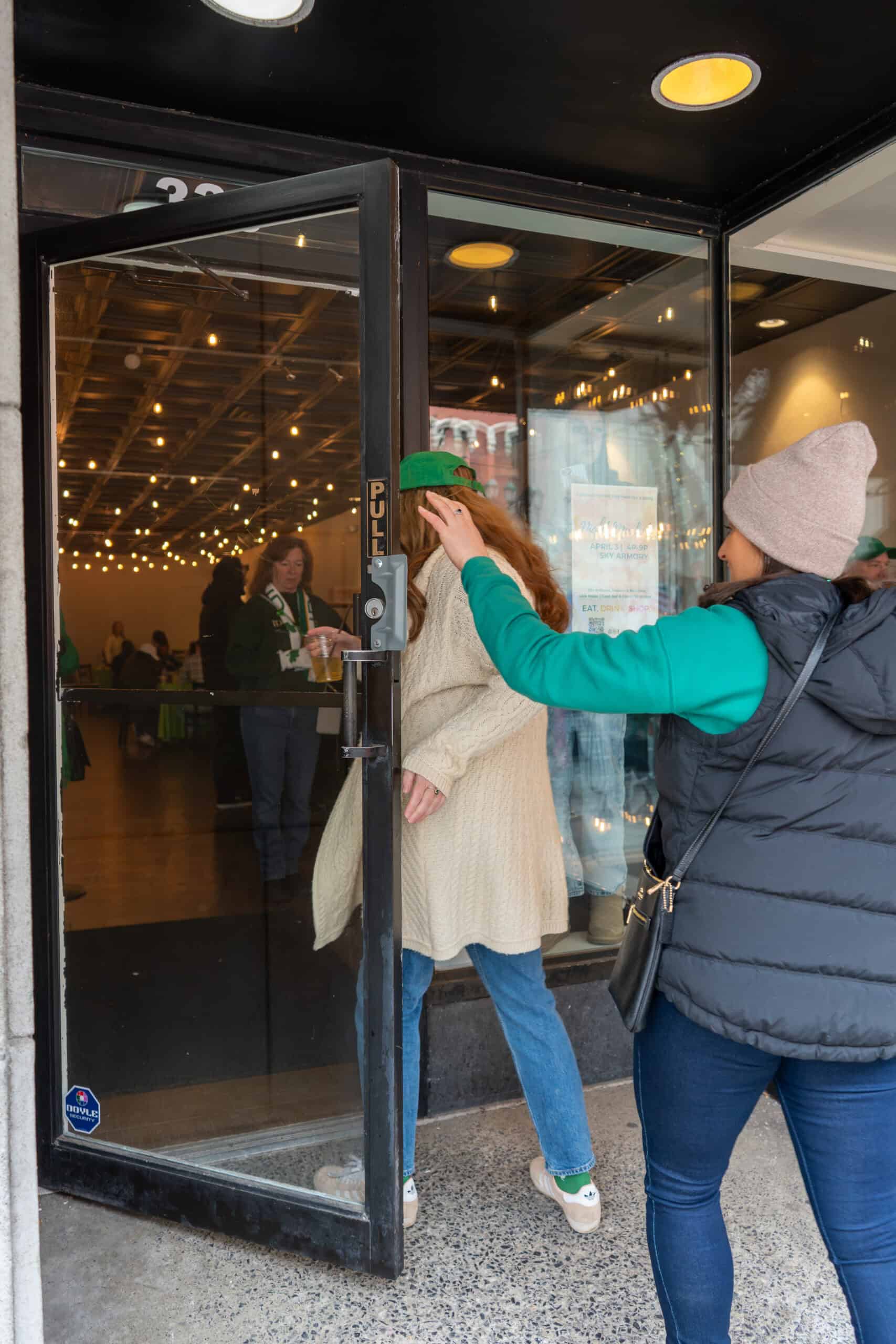Two women entering Sky Armory through glass door in winter attire.