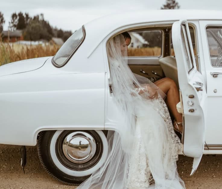Bride in wedding dress sitting in vintage white car with open door.