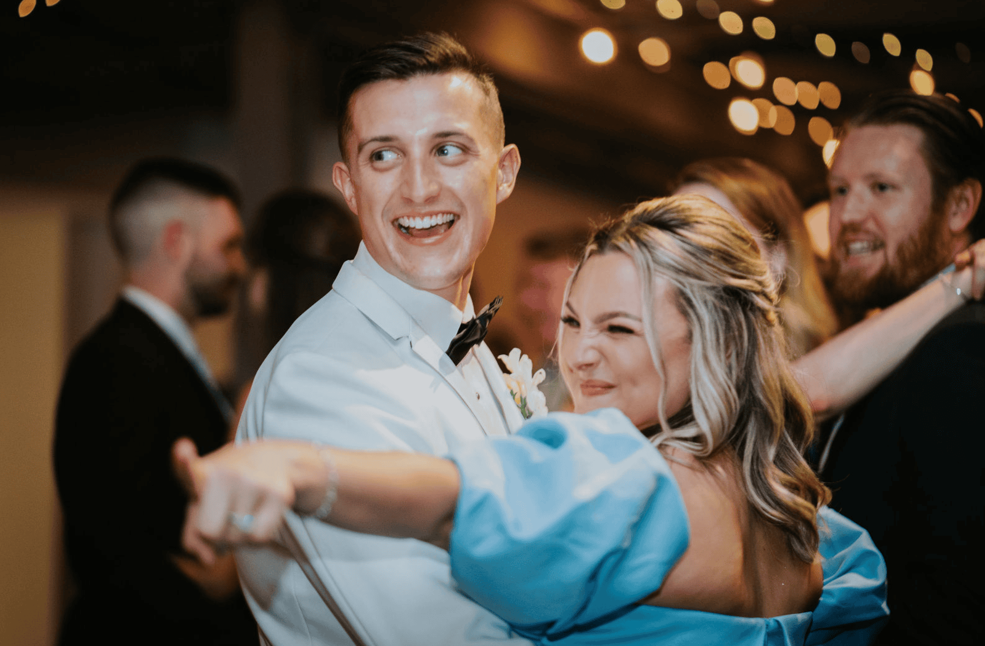 Happy couple dancing at wedding reception with friends and string lights.