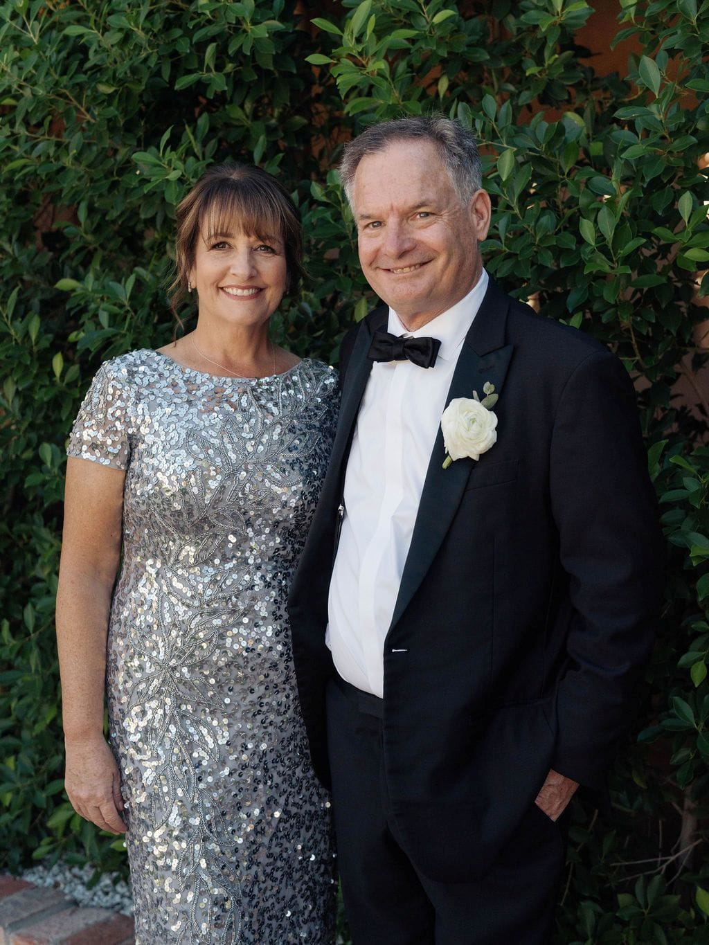 Smiling couple in formal attire outdoors, woman in silver dress, man in tuxedo with boutonniere.