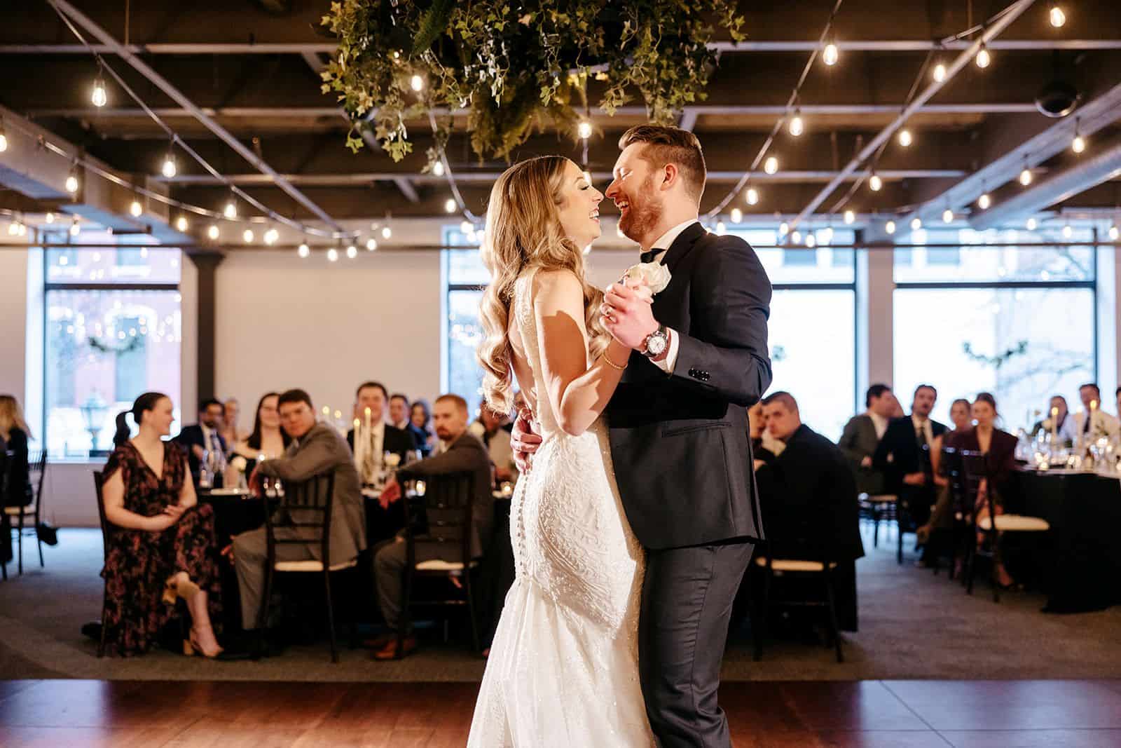 Bride and groom dancing at wedding reception with guests in background.