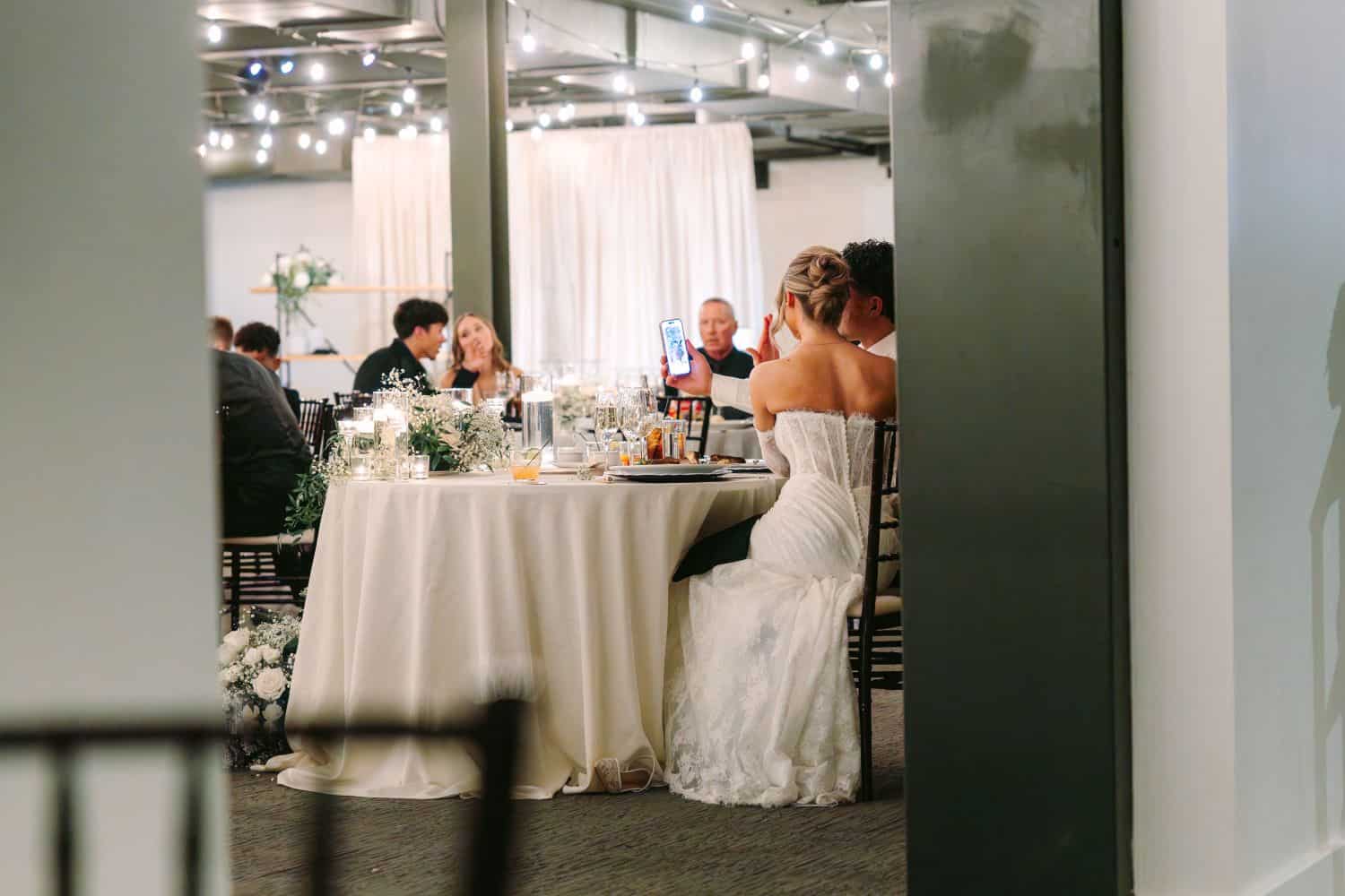 Bride in white wedding dress at reception table with guests and decor.